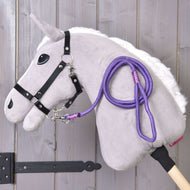 The Hobby Horse LarDen "Hobby Horse - rope stripe Viola" features a plush hobby horse with a white mane, black bridle, purple rope leash, and wooden stick, displayed against a light wooden wall.