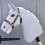 La Hobby Horse LarDen Cavezza con pelliccia - similpelle Walnut è mostrata su un Hobby Horse di peluche grigio con criniera bianca, briglia in similpelle marrone e grandi occhi neri, su un bastone di legno appoggiato a una parete di legno grigia.