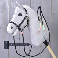 Hobby Horse LarDen "Redini Western Divise Texas" con cavallo in peluche grigio, criniera bianca, briglia nera, redini western divise e nastri colorati al collo, montato su un bastone di legno davanti a una porta di legno grigia.
