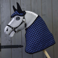 Hobby Horse di Hobby Horse LarDen con criniera bianca e briglia nera che indossa il Set festivo a quadretti blu (cuffia e coperta con motivo pied-de-poule) e posa davanti a una parete in legno grigio.