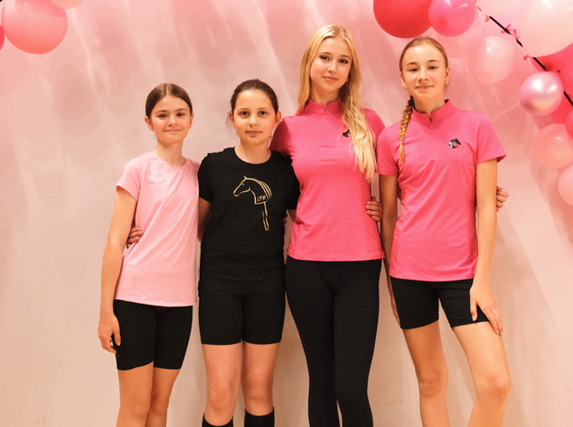 Quattro giovani rider stanno a braccetto davanti a uno sfondo rosa tenue decorato con palloncini rosa, indossando top sportivi rosa e neri con pantaloncini neri o leggings, inclusi dettagli con logo Hobby Horse.
