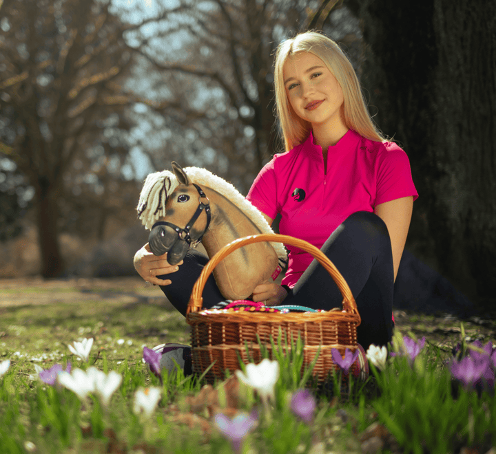 Una giovane donna con una maglietta rosa acceso è seduta sull'erba all'aperto, tiene in mano un Hobby Horse giocattolo e un cestino di vimini pieno di uova colorate, circondata da fiori bianchi e viola in fiore.