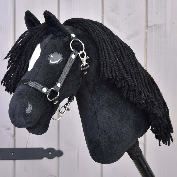 Hobby Horse di peluche nero con lunga criniera in filato, marcature bianche sul muso e briglia nera, montato su un bastone, davanti a uno sfondo in legno chiaro.