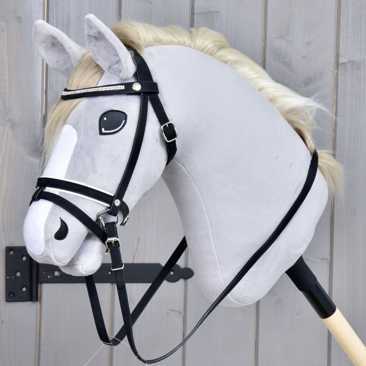 Hobby Horse di peluche con criniera bianca, briglia nera e una striscia bianca sul muso, fissato a un bastone di legno e posizionato davanti a uno sfondo in legno grigio.