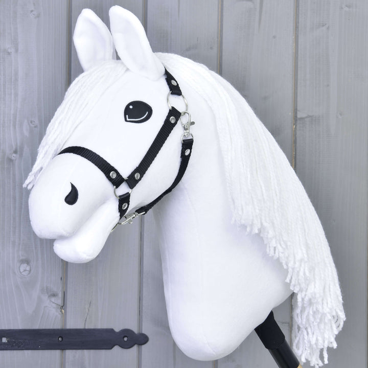 Hobby Horse bianco con briglia nera e lunga criniera bianca in filato, montato su un bastone, su sfondo di parete in legno grigio.