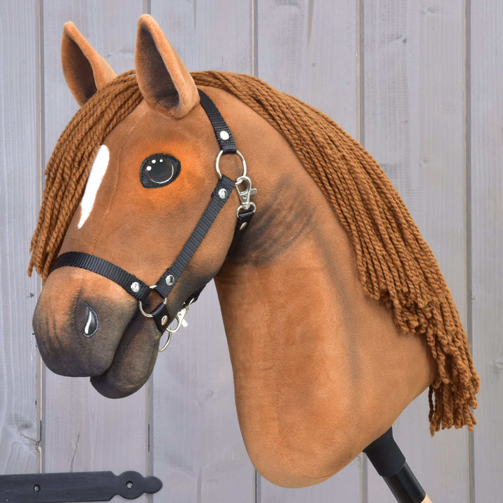A realistic hobby horse with a brown plush head, black bridle, white forehead marking, and long yarn mane, positioned in front of a light wooden wall.