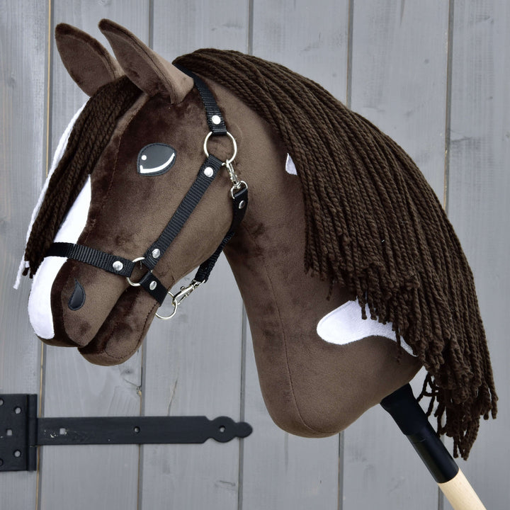 Hobby Horse realistico marrone e bianco con briglia nera e lunga criniera in filato, montato su un bastone di legno, posizionato davanti a una staccionata di legno grigio.