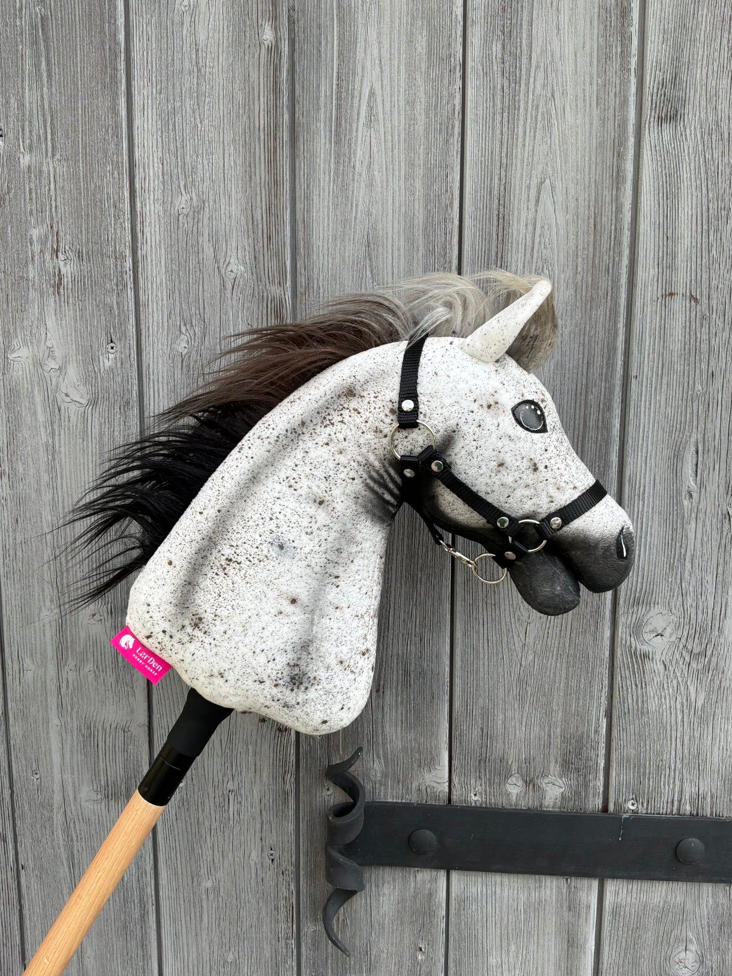 Hobby Horse Luna con capezza nera - taglia S di Hobby Horse LarDen con testa grigia punteggiata, criniera nera e briglia, montato su un bastone di legno e raffigurato appoggiato a una porta rustica in legno con cerniere in metallo.