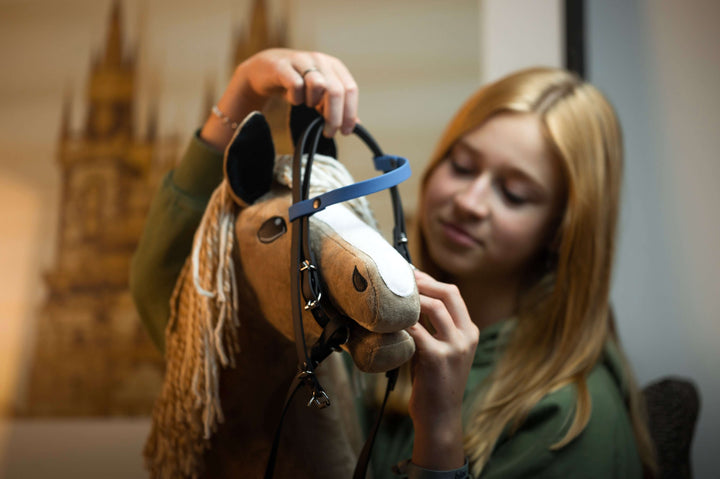 Una giovane ragazza bionda sta mettendo una briglia al suo Hobby Horse, un peluche con criniera beige. È concentrata e sorride leggermente, godendosi l’arte dell’Hobby Horsing in un ambiente interno delicatamente sfocato.