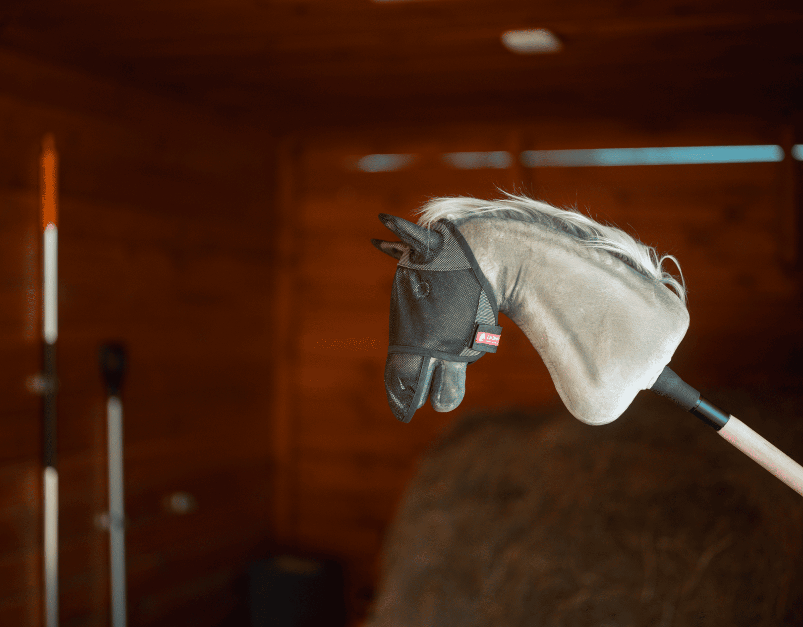 Un Hobby Horse con testa in peluche grigia e briglia nera è montato su un bastone all'interno di una stalla in legno, circondato da attrezzi da fattoria e fieno, ideale per stimolare avventure imaginative di Hobby Horsing.