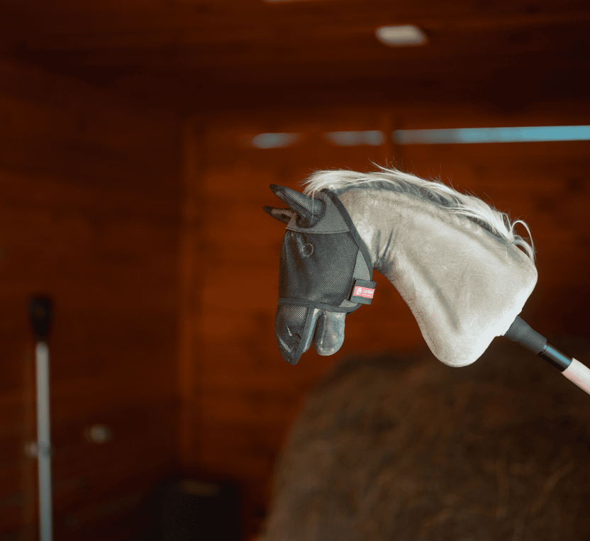 Una testa di Hobby Horse in peluche su un bastone, che indossa una maschera antimosche nera in rete, è mostrata al chiuso con pareti in legno e una pila di fieno, ideale per un divertimento immaginativo con Hobby Horsing.