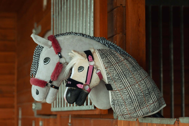 Due hobby horse di peluche con briglie e coperte riposano su una parete divisoria di una stalla in legno, somigliando a veri cavalli in un box. Ideale per gli appassionati di Hobby Horsing, i cavalli bianco e grigio hanno dettagli accurati e finimenti.