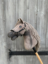 Hobby Horse Swing con cavezza nera - taglia S di Hobby Horse LarDen, con criniera grigia e occhi espressivi, montato su un bastone e appoggiato a una porta di legno invecchiata con venature visibili e ferramenta in ferro.