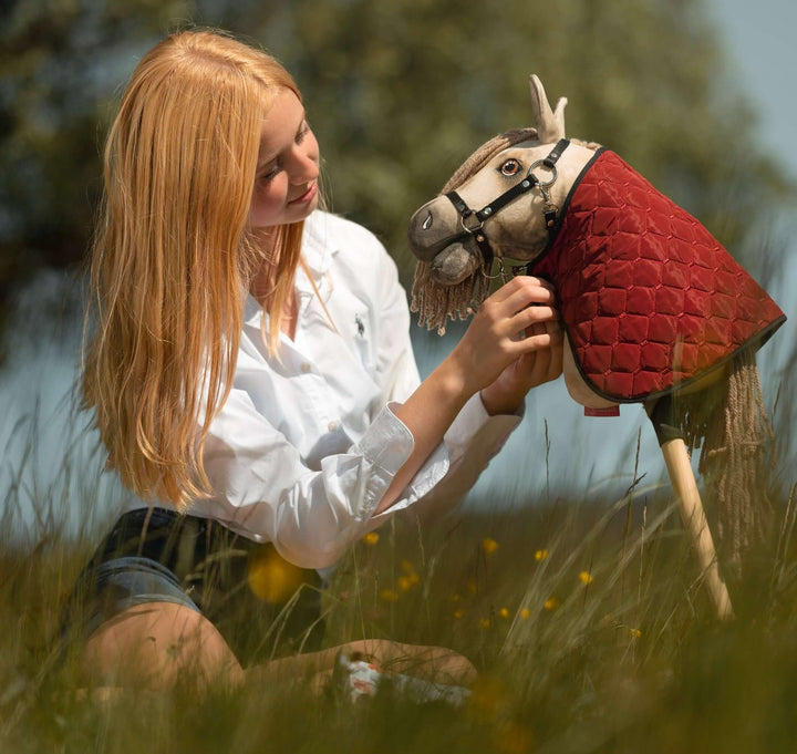 Una giovane persona con lunghi capelli biondi e una camicia bianca è seduta nell’erba alta e sistema delicatamente la briglia di un Hobby Horse con copertura trapuntata rossa, catturando lo spirito fantasioso dell’Hobby Horsing tra il verde sfocato sullo sfondo.