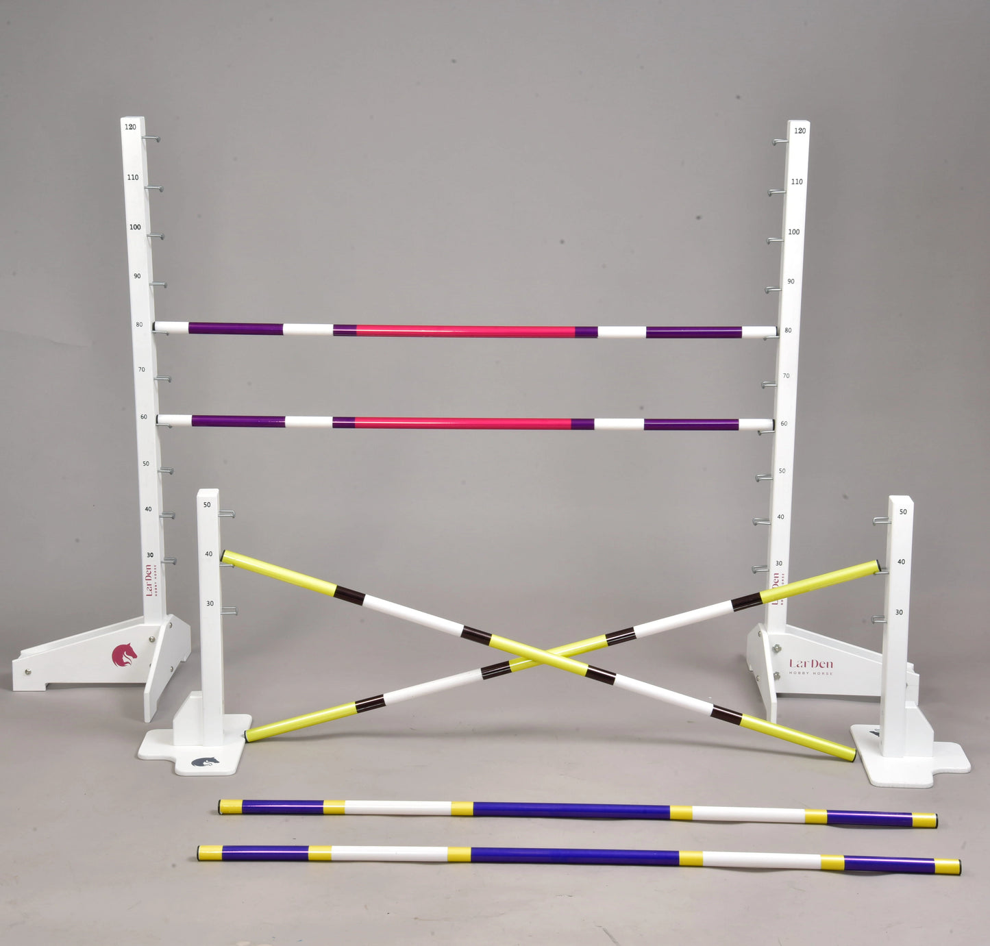 The Hobby Horse LarDen Jump set for Hobby Horsing features adjustable white stands with measuring marks and colored poles in purple, pink, yellow, and black—ideal for jumps or cavaletti, with some poles upright and others on the floor.