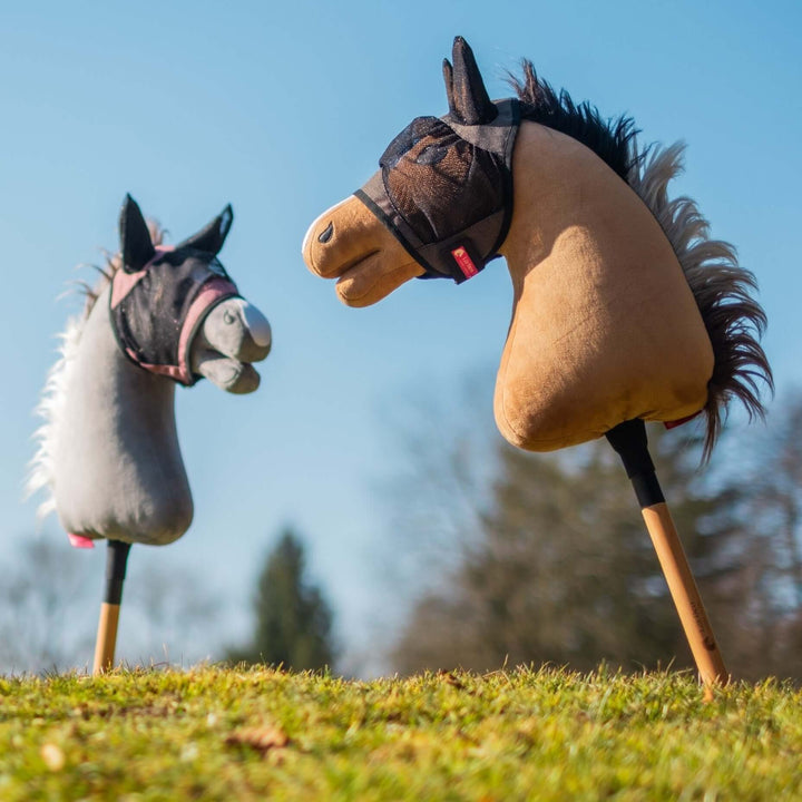 Due hobby horse giocattolo con testa di peluche, uno grigio e uno marrone, stanno in piedi sull’erba contro un cielo azzurro limpido e alberi sfocati. Entrambi i cavalli indossano piccole briglie in tessuto, pronti per imaginative avventure di Hobby Horsing.