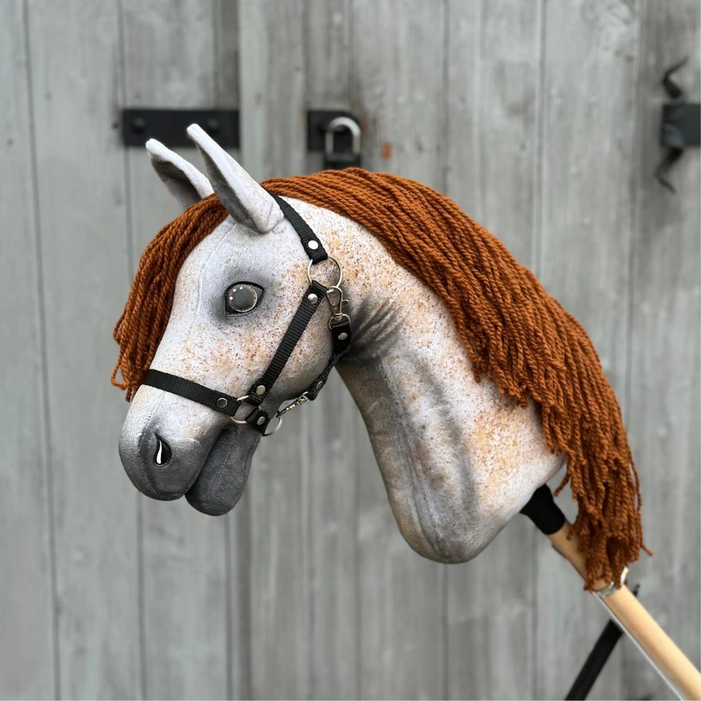 Hobby Horse Pompo con capezza nera di Hobby Horse LarDen con muso grigio maculato, criniera in filato marrone e briglia nera, ispirato allo stallone Hannover, mostrato davanti a una recinzione di legno invecchiata.