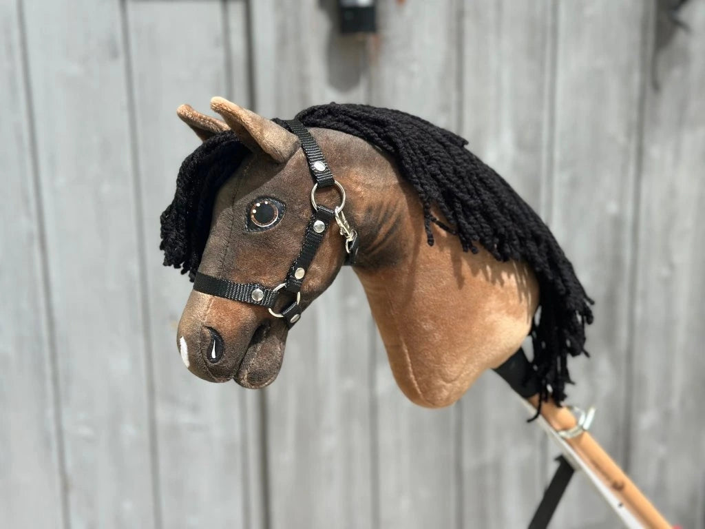 The Hobby Horse LarDen Morgan with Black Halter (size XS) features a brown fabric head, black yarn mane, and black halter on a wooden stick, shown in front of a weathered gray wooden fence.