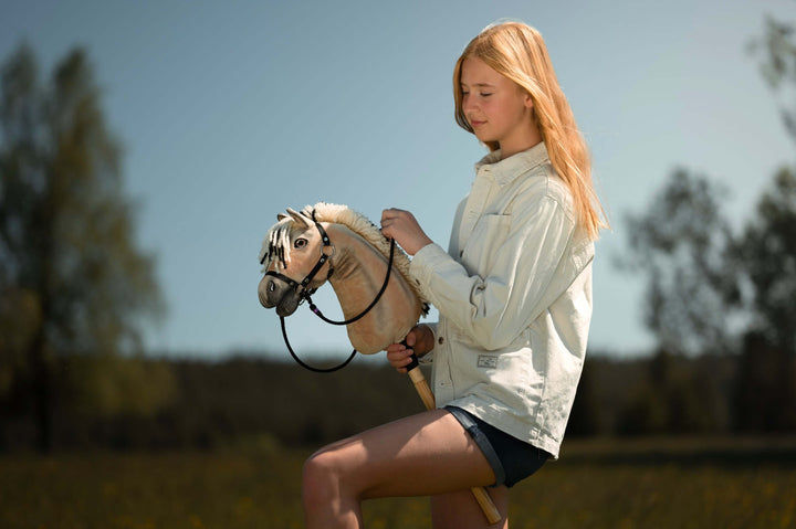 Una giovane persona con lunghi capelli biondi, che indossa una giacca chiara e pantaloncini, pratica Hobby Horsing all’aperto cavalcando un hobby horse in un campo erboso e soleggiato con alberi sullo sfondo.