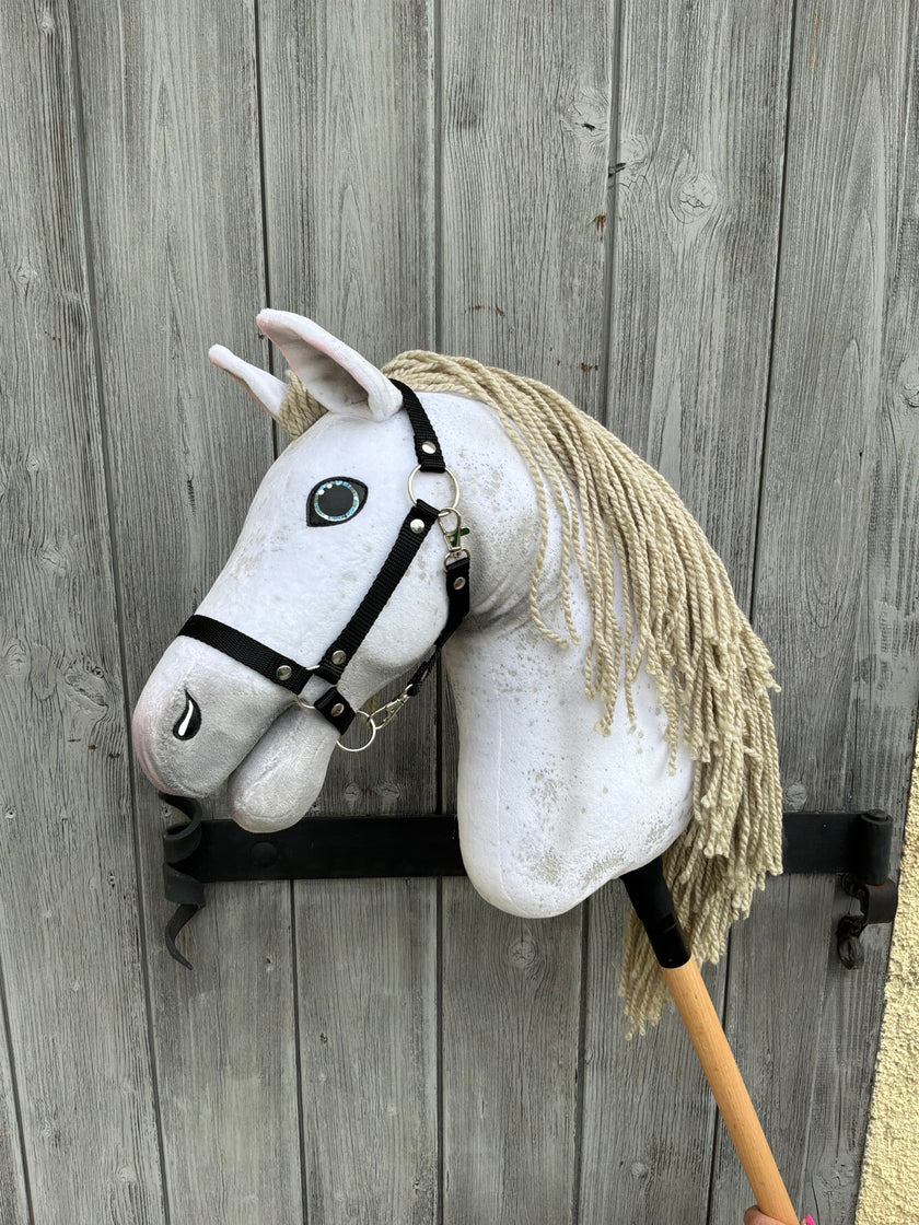 Hobby Horse Victory con capezza nera - taglia M di Hobby Horse LarDen, con mantello bianco e criniera beige in filato, appoggiato sul suo bastone di legno contro una porta di legno grigia invecchiata.