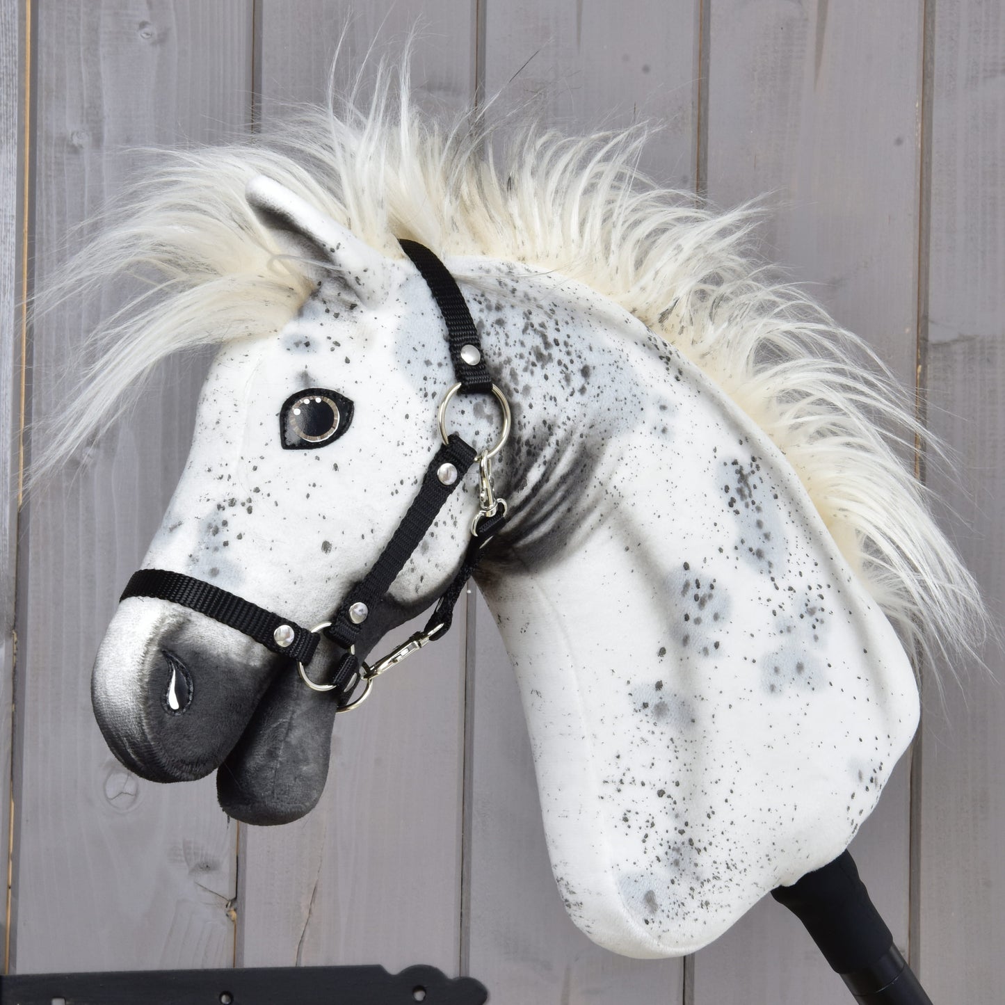 The Hobby Horse LarDen "Hobby Horse Aspen with Black Halter," size S, featuring a white and gray speckled coat reminiscent of an energetic Appaloosa with a fluffy mane and black halter, is positioned in front of a wooden plank wall.