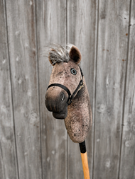 The Hobby Horse LarDen Bolero with Black Halter (size S) features a brown plush head with a gray mane on a wooden stick, shown in front of a weathered fence.