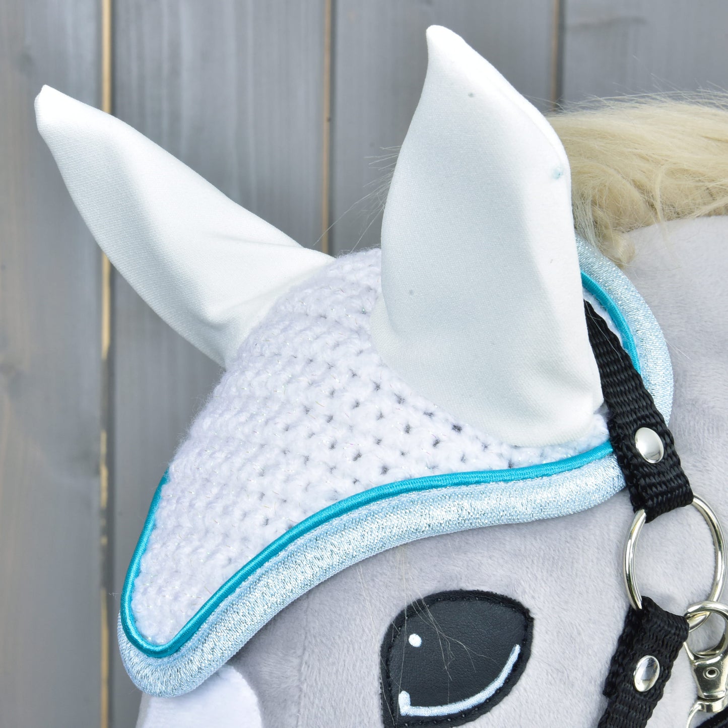 Close-up of a gray stuffed horse toy wearing the Hobby Horse LarDen Ear Bonnet Glitter Cuties, featuring blue trim, two upright white ears, and a black bridle against a light wooden background.