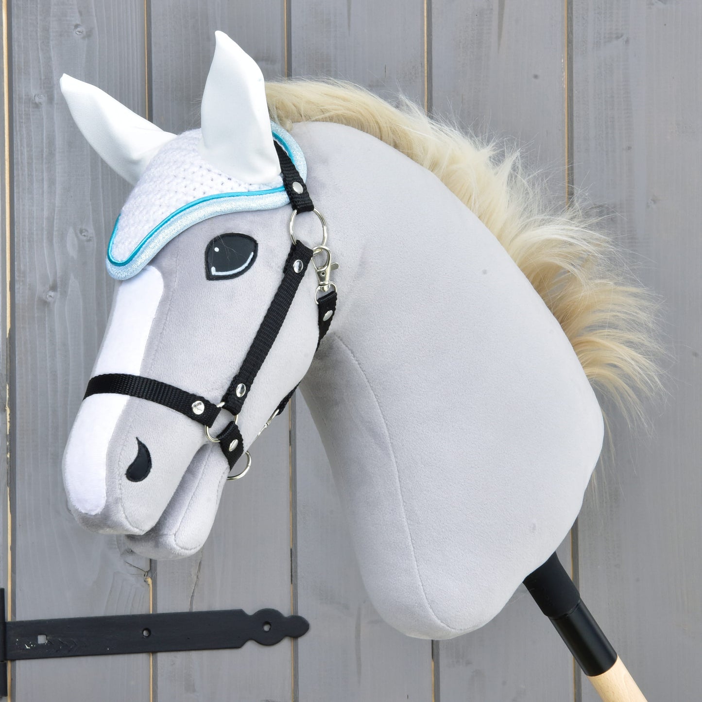 A close-up of a gray plush hobby horse by Hobby Horse LarDen with a white mane, black bridle, and the Ear bonnet Glitter Cuties for fly protection, mounted on a wooden stick in front of a wooden fence.