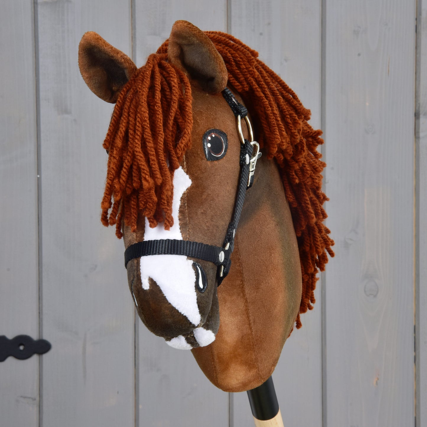 The Hobby Horse LarDen "Cooper with Black Halter" is a brown plush Oldenburg-inspired hobby horse with a thick reddish-brown yarn mane and white facial markings, shown against a gray wooden background.
