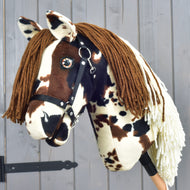 The Hobby Horse LarDen "Hobby Horse Dakota with Black Halter" is a plush hobby horse inspired by the wild mustangs of North America, featuring brown and white spots, a brown yarn mane, and a black halter, posed in front of a gray wooden wall.