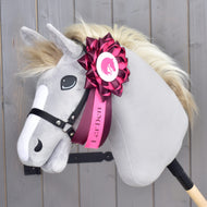 Nastro Hobby Horse Raspberry Rose di Hobby Horse LarDen su un Hobby Horse di peluche con muso grigio, criniera color crema e briglia nera, montato su un bastone e decorato sul collo con un grande nastro da dressage rosa e bianco con logo LarDen. Sfondo in pannelli di legno.