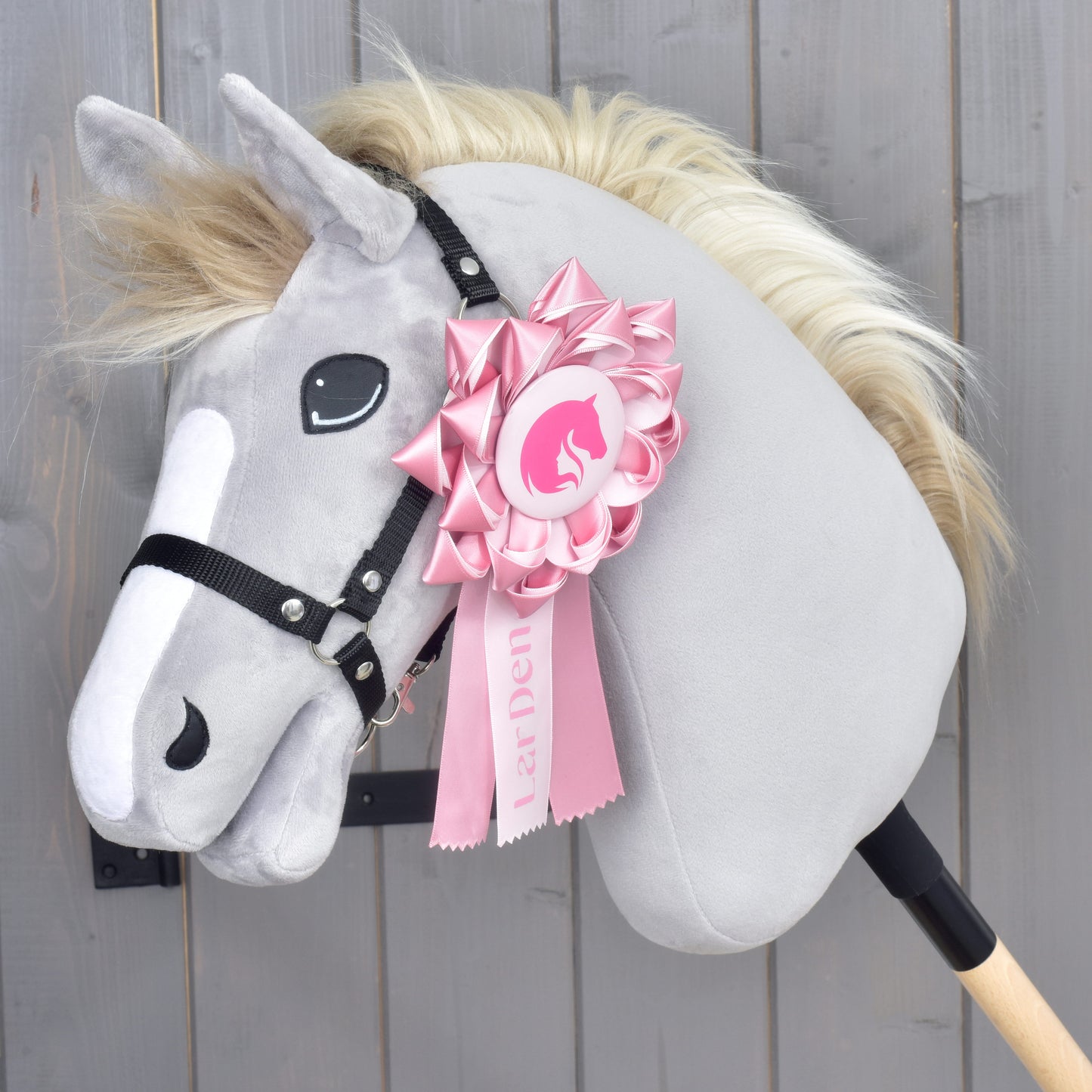 Hobby Horse LarDen Nastro Hobby Horse LarDen Rosa con cavallo di peluche grigio, briglia nera e criniera color crema, decorato con un grande nastro da concorso rosa sulla testa, su sfondo in legno.