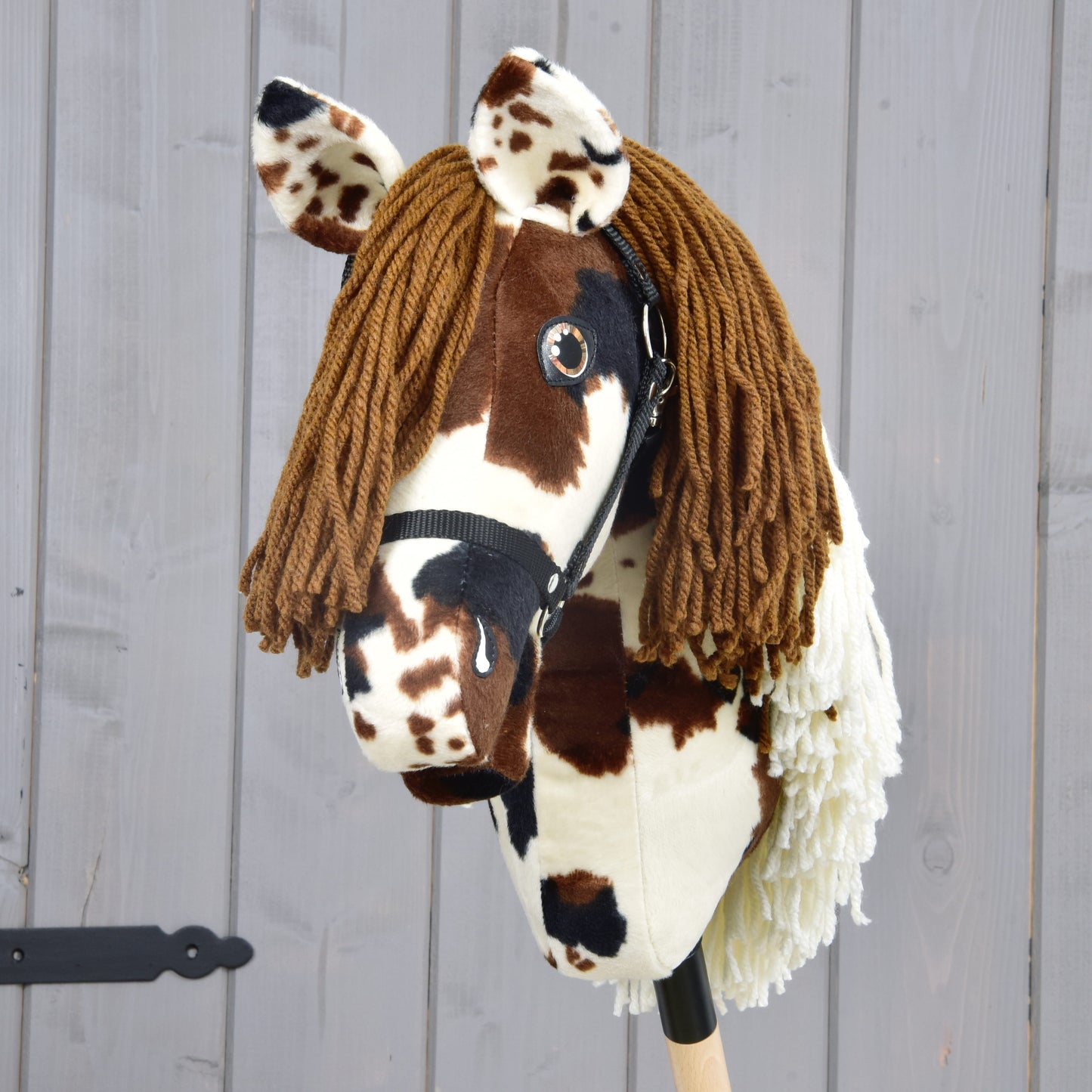 The Hobby Horse Dakota with Black Halter by Hobby Horse LarDen features a brown and white plush head, brown yarn mane, and black halter on a stick, photographed against a gray wooden background. Suitable for aspiring riders of any gender.