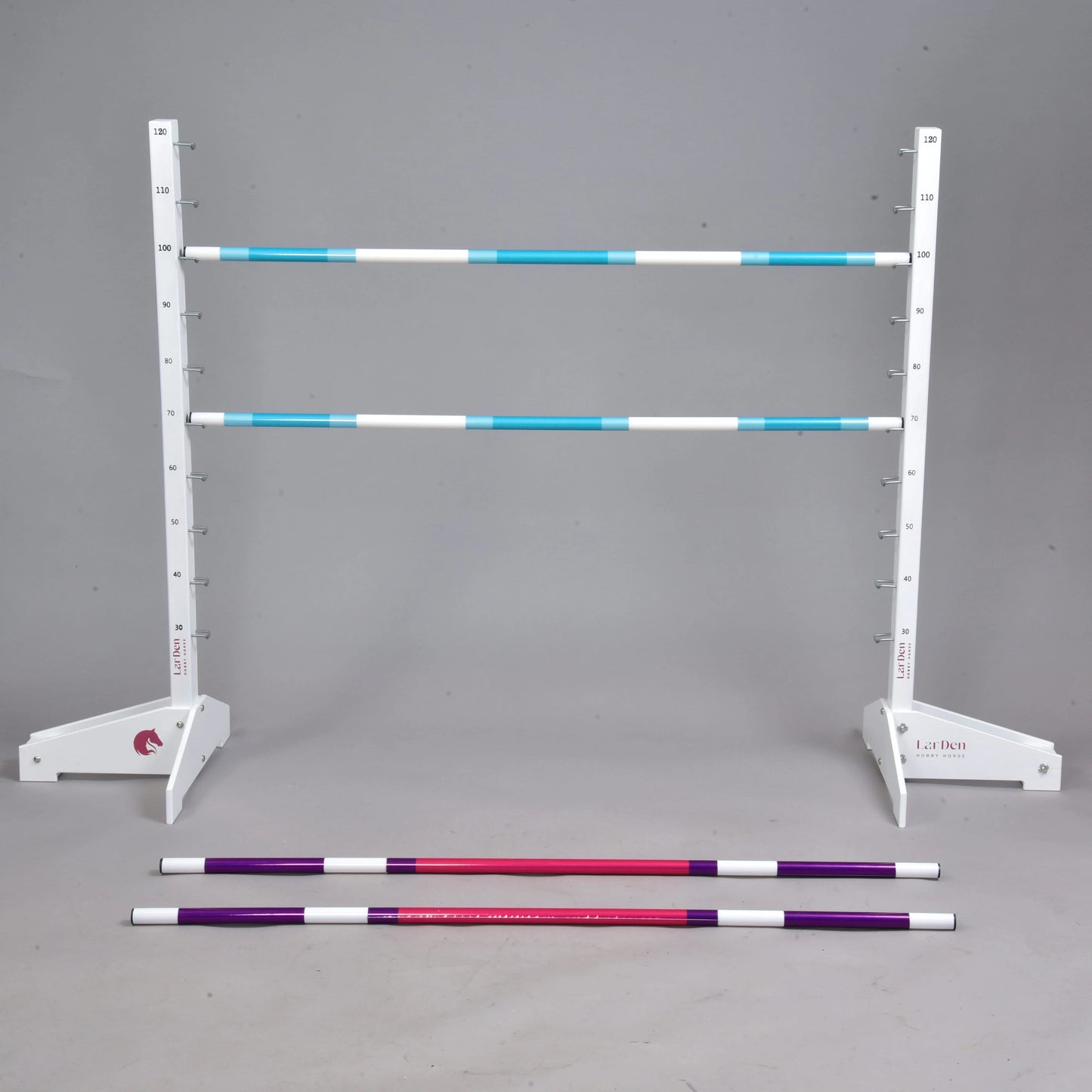 The Hobby Horse LarDen Jump 120 cm / 47 inch with Extra Poles set features a white adjustable stand holding two blue and white striped rails, plus red and purple poles on a grey floor. Perfect for Hobby Horsing, with a plain grey background.