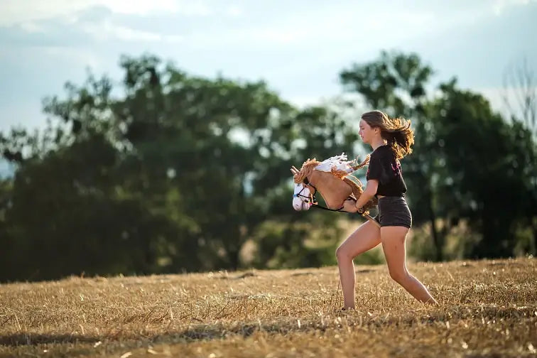 Una giovane persona corre attraverso un campo erboso e secco praticando Hobby Horsing con un Hobby Horse, con alberi e cielo azzurro sullo sfondo.