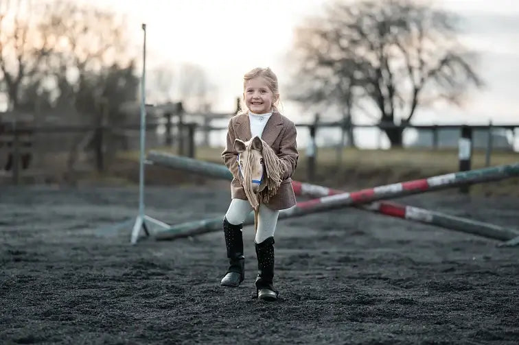 Una giovane ragazza sorride mentre si diverte con Hobby Horsing, fingendo di cavalcare un Hobby Horse all'aperto su un campo in terra battuta con pali incrociati e alberi sullo sfondo. Indossa una giacca marrone, pantaloni bianchi e stivali da equitazione neri.
