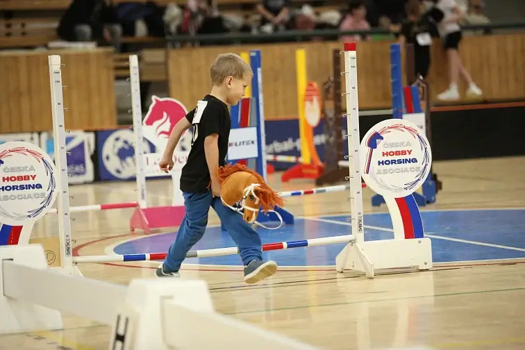 Un giovane ragazzo partecipa a una competizione di Hobby Horse, camminando con un cavallo giocattolo sul pavimento di una palestra, circondato da ostacoli colorati per il salto e cartelli dell'evento di Hobby Horsing.