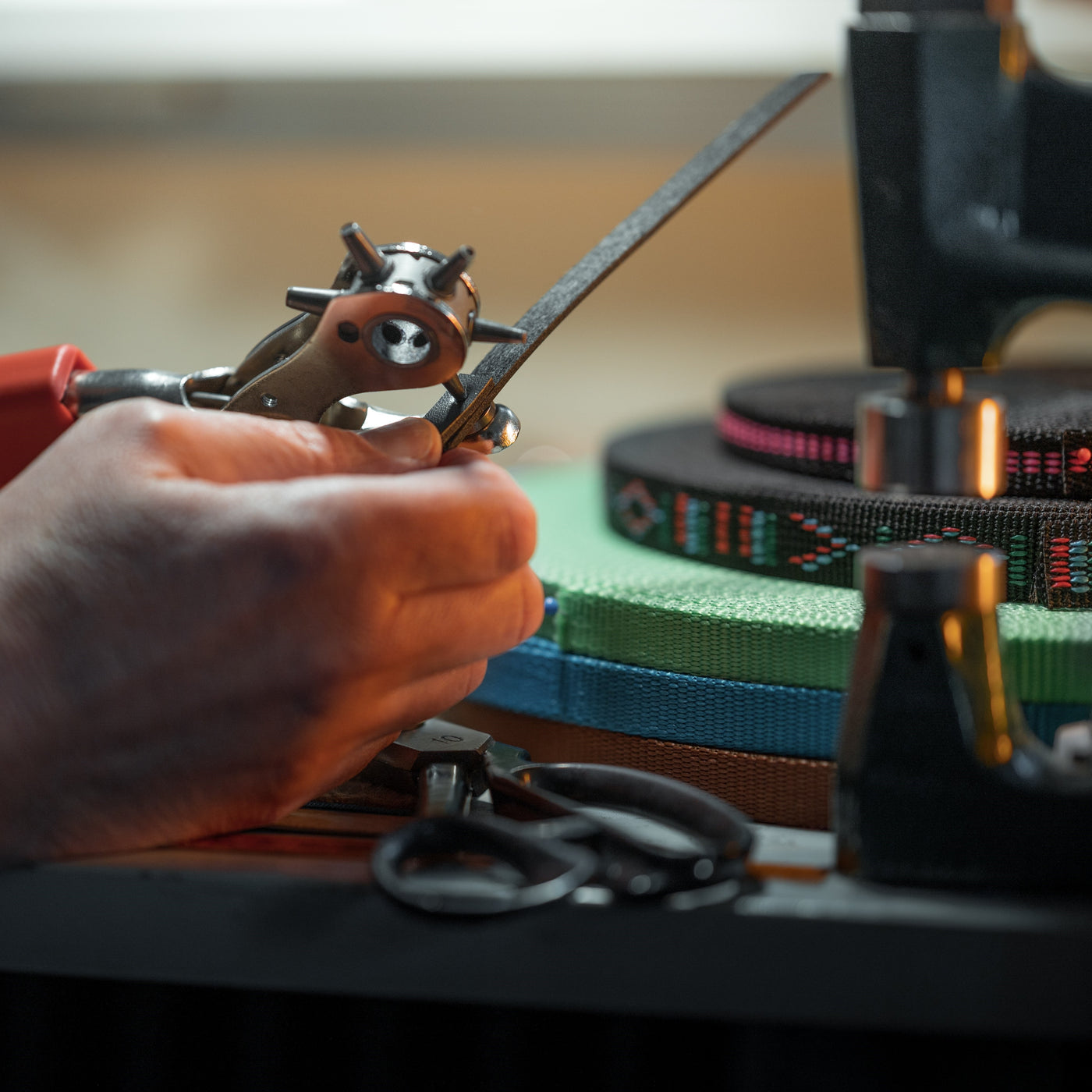 A close-up of a person's hands using a leather hole punch tool on a strip of black leather, possibly crafting gear for Hobby Horsing, with colorful woven bands and a pair of scissors on a table in the background.