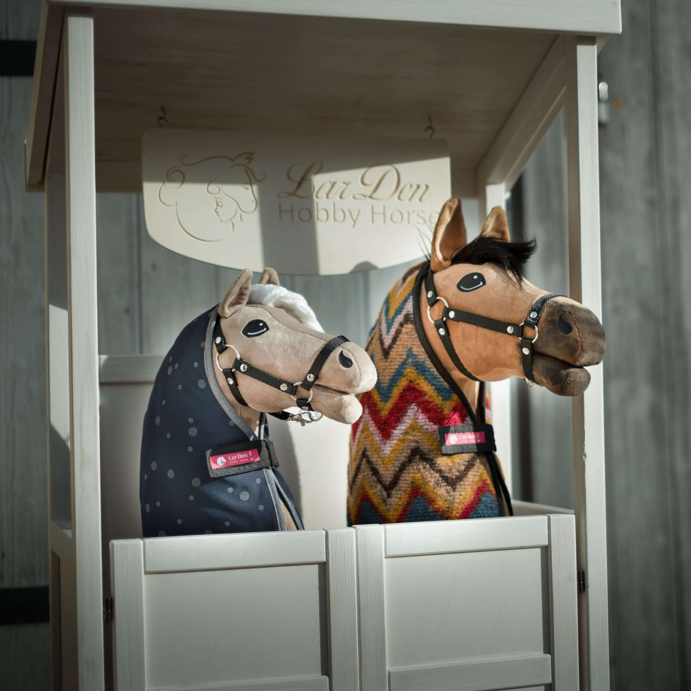 Due Hobby Horse di peluche con briglie e coperte, uno con coperta blu a pois e l'altro con fantasia zigzag colorata, sono esposti affiancati in una cabina di legno bianca con la scritta LarsDen Hobby Horsing.