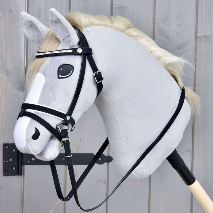 Hobby Horse LarDen Briglia Completa De Luxe con 2 frontalini su realistico Hobby Horse grigio con criniera bianca e capezzina combinata, montato su bastone di legno davanti a uno sfondo in legno grigio.