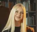 Una giovane donna con lunghi capelli biondi lisci sorride alla fotocamera. Indossa un top nero, con una libreria sfocata sullo sfondo. Una mano le poggia delicatamente sulla spalla, suggerendo un momento affettuoso dopo una divertente giornata di Hobby Horsing.