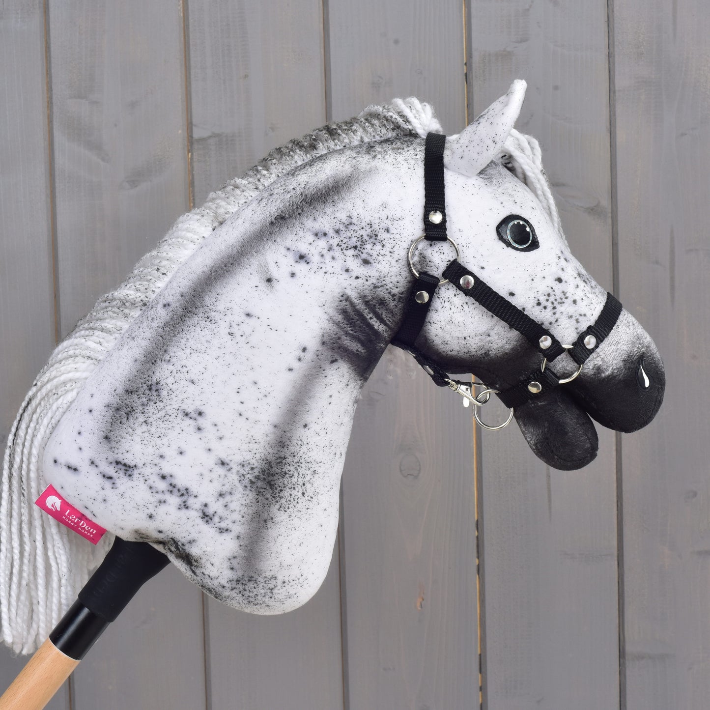The Hobby Horse LarDen's Hobby Horse Naivy with Black Halter - size S, featuring a striking white and grey speckled design, stands proudly mounted on a wooden stick in front of a gray wooden wall.