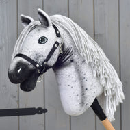 The Hobby Horse LarDen "Hobby Horse Naivy with Black Halter" (size S), featuring a gray and white Appaloosa design with a black halter and yarn mane, is mounted on a wooden stick and set against a gray wooden wall.