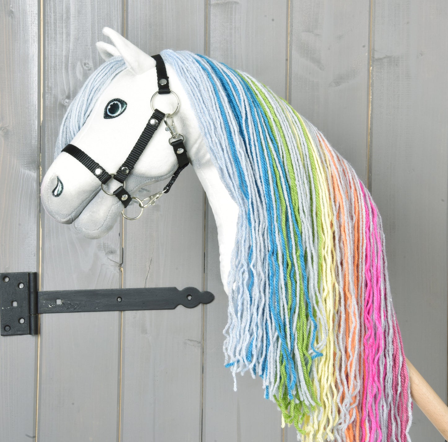The Hobby Horse LarDen "Hobby Horse Rainbow New" features a white face, black bridle, and long rainbow-colored yarn mane, standing in front of a grey wooden door.