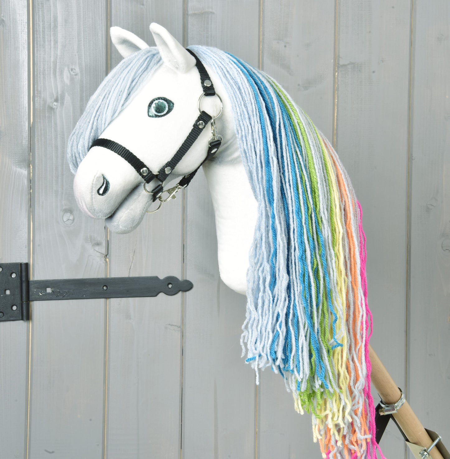 The Hobby Horse Rainbow New by Hobby Horse LarDen features a handmade white face, blue eyes, black bridle, and rainbow mane with blue, green, yellow, and pink yarn against a light gray wooden background for a fairy-tale touch.