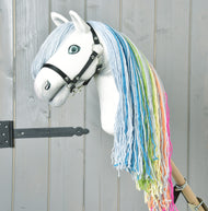 The Hobby Horse Rainbow New by Hobby Horse LarDen features a handmade white face, blue eyes, black bridle, and rainbow mane with blue, green, yellow, and pink yarn against a light gray wooden background for a fairy-tale touch.