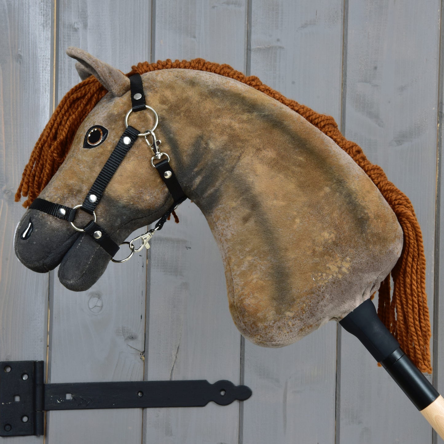 A close-up of the Hobby Horse LarDen Rafaello with Black Halter (size S), featuring a brown body, yarn mane, button eyes, black bridle and reins, and a beige wooden stick, set against a gray wooden wall background.
