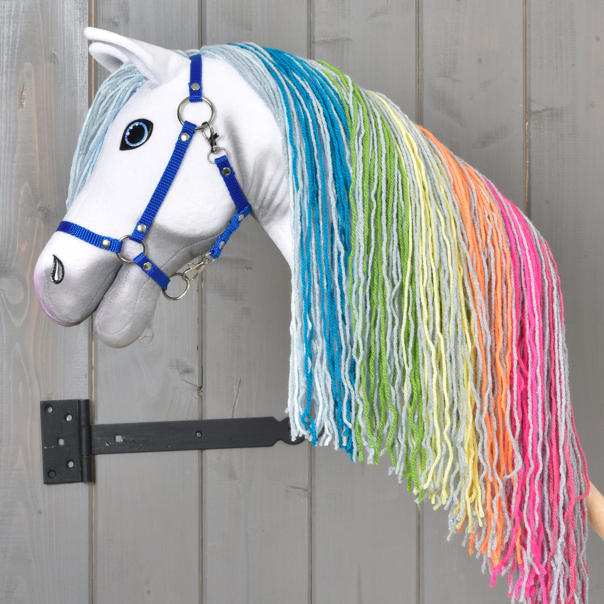 A Hobby Horse LarDen “Hobby Horse Rainbow New” with a blue bridle and a long, rainbow yarn mane hangs on a wall-mounted holder against a wooden background, adding fairy-tale charm to the room.
