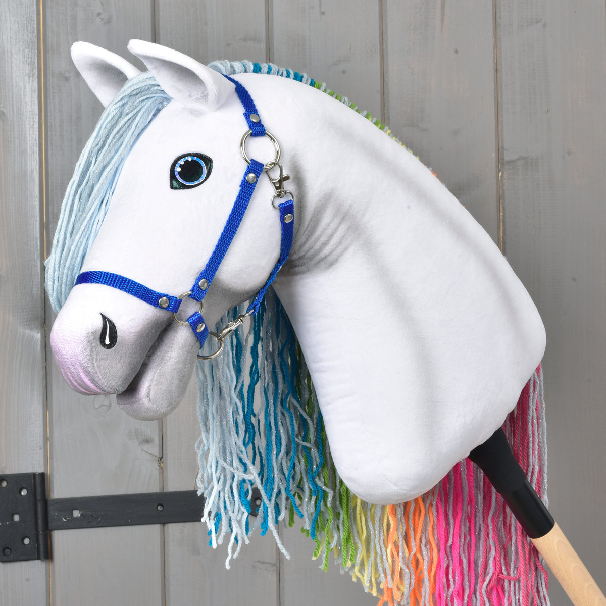 A whimsical Hobby Horse Rainbow New by Hobby Horse LarDen, featuring blue reins, a light blue yarn mane, and a fairy-tale rainbow mane for its tail, set against a gray wooden background.