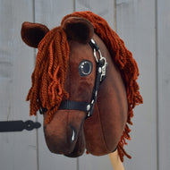 A brown fabric Hobby Horse Rebel with Black Halter - size XS from Hobby Horse LarDen, designed to resemble a Hanoverian stallion with an orange yarn mane, is displayed in front of a gray wooden wall.